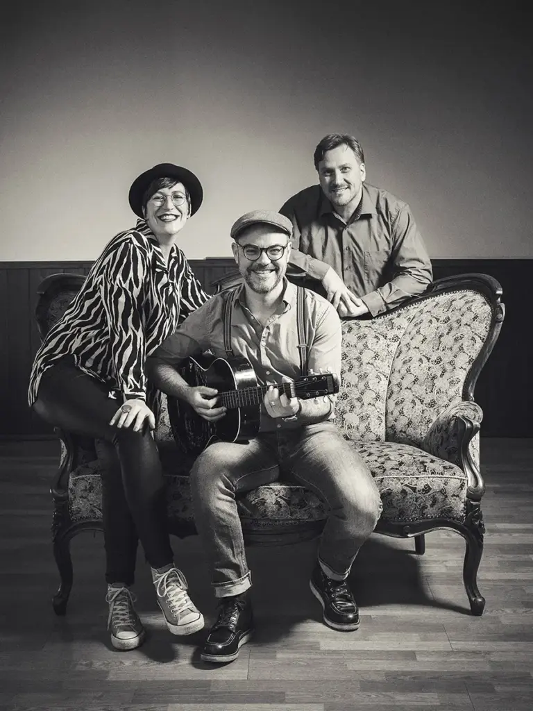 Schwarz-Weiß-Foto: Conny Morath mit Hut, Stephan Schmitt mit Gitarre und Thomas Koch lachen gemeinsam auf einem Vintage-Sofa